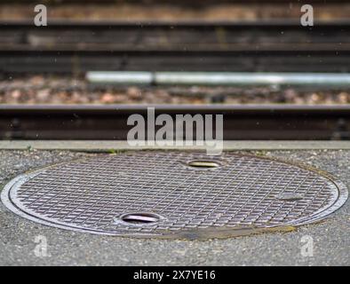 Nahaufnahme Nahaufnahme der Mannlochabdeckung auf nassem Gehweg mit Eisenbahngleisen im Hintergrund, industrielle urbane Szene. Im Hintergrund: Industrielle urbane Szene. Stockfoto