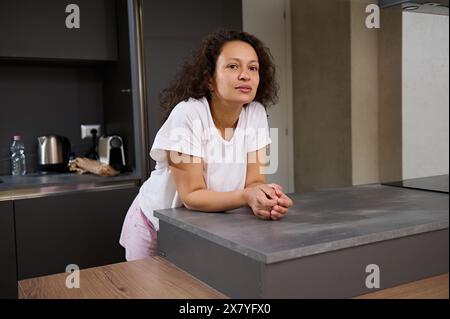 Porträt einer nachdenklichen Frau in der Küche, während sie morgens aufwacht, in weißen Pyjamas gekleidet, an der Küchentheke steht und traumhafte Looki sieht Stockfoto