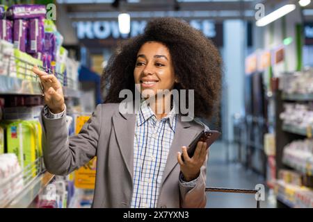 In einem Lebensmittelgeschäft kauft eine Frau mit lockigen Haaren und einer formellen Jacke mit einem Smartphone ein, zeigt auf Produkte in einem Regal, zeigt moderne Einkaufsgewohnheiten und den Einsatz von Technologie im Einzelhandel Stockfoto