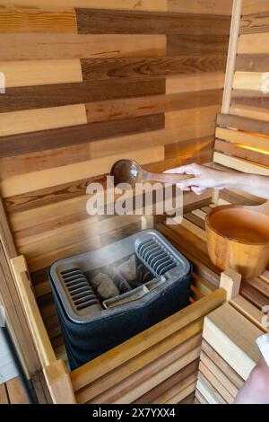 Frau, die Wasser auf heißen Stein in der Sauna gießt. Dampf aus Wasser auf den Steinen, Spa und Wellness-Konzept Stockfoto