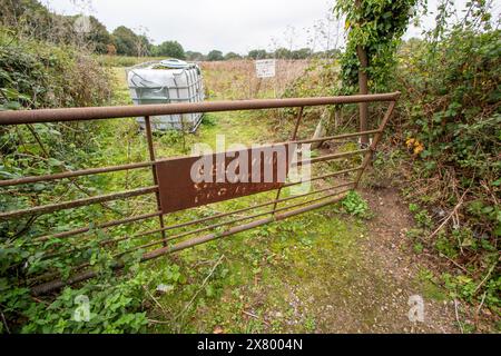 Ein rostendes 5-Bar-Tor im ländlichen Essex mit einem fast unleserlichen Hinweis schützt einen riesigen Plastiktank, der auf dem Feld dahinter steht. Sorgfältige Haltung. Stockfoto