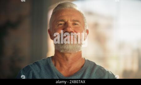 Porträt eines glücklichen lächelnden Mannes mittleren Alters, der in die Kamera schaut und sich anstrengt, Eine schwere Hantel zu heben. Senior männlich, die zu Hause trainieren. Wellness- und Fitnesskonzept. Stockfoto
