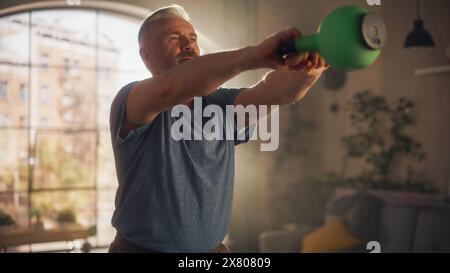 Starker Athletic Fit Senior man hebt und schwingt eine schwere Kettlebell, macht Übungen zur Stärkung des Kerns während des morgendlichen Workouts zu Hause in der Sunny Apartment. Konzept von Gesundheit und Fitness. Stockfoto