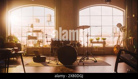 Establishment Shot: Musikprobe Studio im Loft Room mit Drum Set in der Mitte. Stilvolles Interieur mit zwei großen Fenstern, gemütlichem Sofa, Regalen und Pflanzen. Sonniger, heller Tag und Blick auf die Stadt. Stockfoto