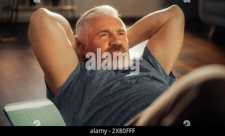 Nahaufnahme Porträt eines glücklichen Seniorenmannes, der am sonnigen Morgen zu Hause trainiert. Mann mittleren Alters, der auf dem Boden liegt und Übungen zur Stärkung des Kerns macht. Wellness und Wohlbefinden Lifestyle. Stockfoto