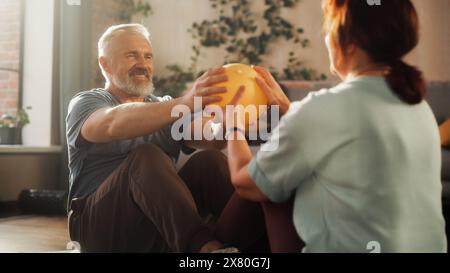 Seniorenpaar, das im sonnigen Wohnzimmer gemeinsam Übungen zur Stärkung des Kerns und Crunches-Workout macht. Glückliche Männer und Frauen mittleren Alters motivieren sich gegenseitig, gesund zu sein. Stockfoto