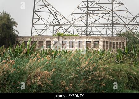 Verlassenes Industriebauhaus am Fuße eines elektrischen Pylons am Ufer des Nils mit Vegetation im Vordergrund. Stockfoto