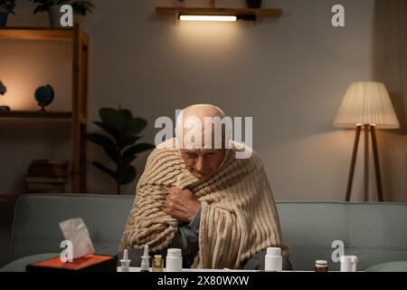 Ungesunder Mann, der mit Decke bedeckt sitzt, sich krank fühlt, an Grippe leidet. Ungesunder älterer Mann, der Grippe oder Erkältung zu Hause bekämpft. Stockfoto