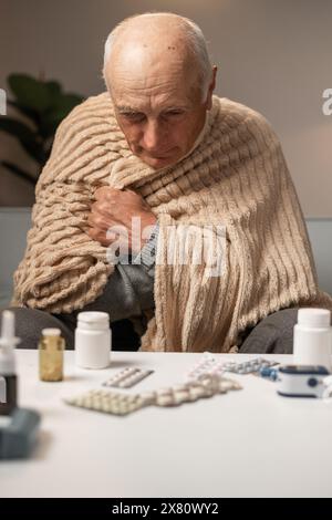 Kranker älterer Mann, der sich kalt fühlt, mit einer Decke bedeckt, auf dem Sofa sitzt, kranker Mann, der vor Kälte zittert, sich zu Hause aufwärmt. Behandlung der Influenza Stockfoto