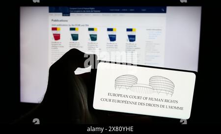 Person, die ein Mobiltelefon mit dem Logo des Internationalen Gerichtshofs für Menschenrechte (EMRK) vor der Webseite hält. Schwerpunkt auf dem Display des Telefons. Stockfoto