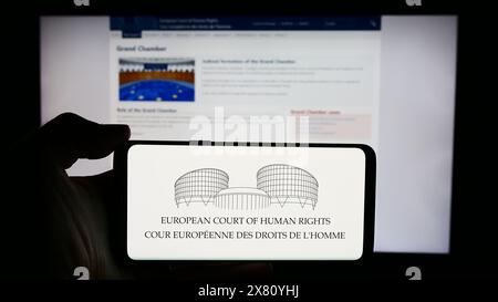 Person, die ein Mobiltelefon mit dem Logo des Internationalen Gerichtshofs für Menschenrechte (EMRK) vor der Webseite hält. Schwerpunkt auf dem Display des Telefons. Stockfoto