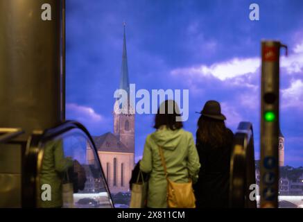 Zürich, Schweiz - 16. Mai 2024: Zwei Individuen auf der Rolltreppe, vor städtischer Sicht auf Zürich mit historischer Kirche und dramatischem Himmel. Stockfoto