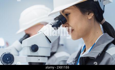 Nahaufnahme Porträt eines jungen asiatischen Industriewissenschaftlers mit weißem Schutzhelm, der an einem Schreibtisch in einer Fabrik arbeitet und die Fertigungsteile mithilfe eines Mikroskops auf Qualität untersucht. Stockfoto