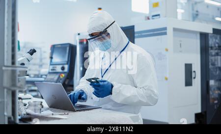 Modernes Technologiekonzept: Ingenieur im Overall, der an Laptop-Computern arbeitet und eine Leiterplatte mit Microchips untersucht. Techniker In Der Elektronikfabrik Testet Neue Elektrische Geräte. Stockfoto