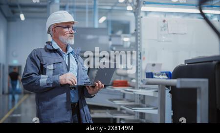 Bärtiger Chefingenieur mittleren Alters, der in einer Fabrik spaziert, einen Arbeitsanzug, eine stilvolle blaue Brille und einen weißen Schutzhelm trägt. Spezialist für Schwerindustrie, der an Laptops arbeitet. Stockfoto