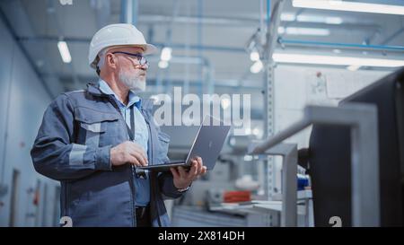 Porträt eines bärtigen General Managers mittleren Alters, der in einer Fabrik steht, einen Arbeitsanzug, eine stilvolle blaue Brille und einen weißen Schutzhelm trägt. Spezialist für Schwerindustrie, der an Laptops arbeitet. Stockfoto