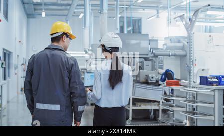 Rückansicht: Zwei Mitarbeiter der Schwerindustrie in Sicherheitsuniformen und Hart-Hüten, die in einer Fabrik laufen und sprechen. Zwei Ingenieure suchen nach Brainstorming und lösen Probleme Stockfoto