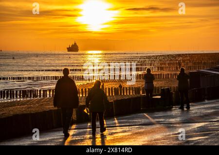 Sonnenuntergang am Strand von Zoutelande, Strand mit Holzpfahlbrüchen, Touristen, Frachtschiff Segeln in Richtung Westerschelde, Zeeland, Niederlande Stockfoto