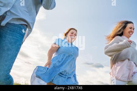 Porträt von drei fröhlich lächelnden Frauen, die auf der Wiese rennen. Niedrigwinkelfoto am blauen Himmel mit Wolken. Freundschaft der Frau, Beziehungen und Stockfoto