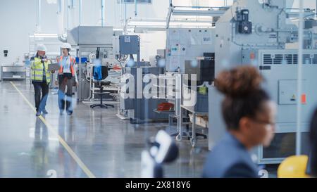 Zwei professionelle Spezialisten für Schwerindustrie für Sicherheitsuniform und Schutzhüte, die in einer Fabrik laufen und sprechen. Afroamerikanischer Ingenieur, der im Vordergrund arbeitet Stockfoto