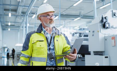 Porträt eines bärtigen Ingenieurs mittleren Alters, der in einer Fabrik steht und eine Warnjacke und einen weißen Schutzhelm trägt. Spezialist für Schwerindustrie, der an Laptops arbeitet. Stockfoto