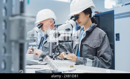 Electronic Manufacturing Factory: Zwei Ingenieure untersuchen kleine Teile von Industrieanlagen mit einem Mikroskop. Techniker testen neue Werkskomponenten auf Produktionsqualität. Stockfoto