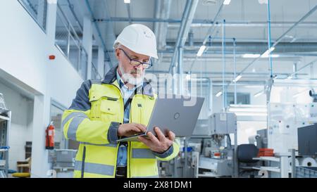 Porträt eines bärtigen Ingenieurs mittleren Alters, der in einer Fabrik steht und eine Warnjacke und einen weißen Schutzhelm trägt. Spezialist für Schwerindustrie, der an Laptops arbeitet. Stockfoto