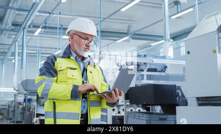 Porträt eines bärtigen Ingenieurs mittleren Alters, der in einer Fabrik steht und eine Warnjacke und einen weißen Schutzhelm trägt. Spezialist für Schwerindustrie, der an Laptops arbeitet. Stockfoto