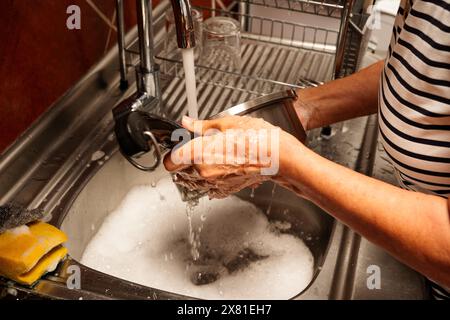 Eine Frau wäscht ein schmutziges Geschirr in einem Waschbecken. Das Waschbecken ist voll mit Seifenwasser und die Frau schrubbt die Schale mit ihren Händen Stockfoto