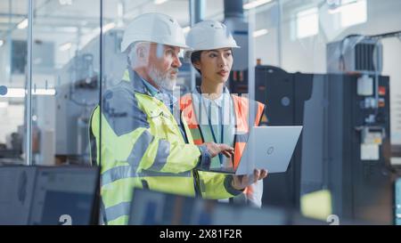 Zwei professionelle Mitarbeiter der Schwerindustrie, die in der Fabrik Schutzhüte tragen. Überprüfung und Besprechung der Arbeiten am Laptop-Computer. Asiatischer Qualitätskontrolleur und Techniker bei der Arbeit. Stockfoto