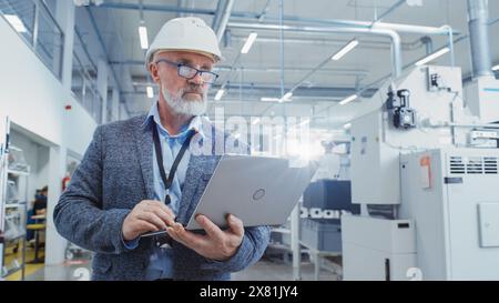 Porträt eines bärtigen General Managers mittleren Alters, der in einer Fabrik spaziert, in lässigem Anzug und einem weißen Schutzhelm. Spezialist für Schwerindustrie, der an Laptops arbeitet. Stockfoto