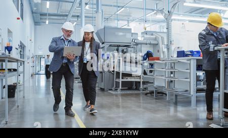 Zwei professionelle Mitarbeiter der Schwerindustrie, die in der Fabrik Schutzhüte tragen. Gehen Sie und diskutieren Sie über industrielle Maschinenanlagen, arbeiten Sie an Laptops. Afroamerikanischer Ingenieur und Techniker bei der Arbeit. Stockfoto