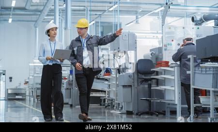 Zwei junge asiatische Ingenieure für Schwerindustrie in Hart-Hüten, die mit Laptop-Computer laufen und in einer Fabrik reden. Zwei Fertigungsmitarbeiter arbeiten in der Produktionsanlage. Stockfoto