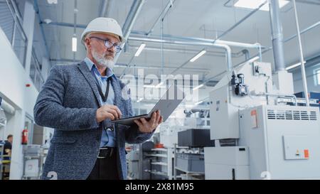 Porträt eines bärtigen Chefingenieurs mittleren Alters, der in einer Fabrikhalle steht und einen lässigen Anzug und einen weißen Schutzhelm trägt. Spezialist für Schwerindustrie, der an Laptops arbeitet. Stockfoto