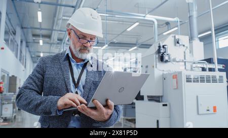 Porträt eines bärtigen Chefingenieurs mittleren Alters, der in einer Fabrikhalle spaziert, in lässigem Anzug und einem weißen Schutzhelm. Spezialist für Schwerindustrie, der an Laptops arbeitet. Stockfoto