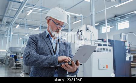 Porträt eines bärtigen General Managers mittleren Alters, der in einer Fabrikhalle steht, in lässigem Anzug und einem weißen Schutzhelm. Spezialist für Schwerindustrie, der an Laptops arbeitet. Stockfoto