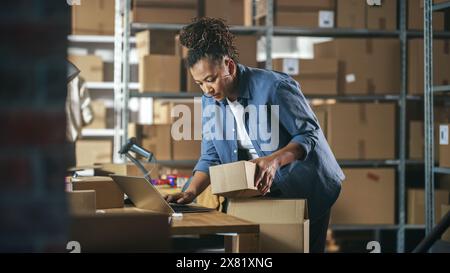 Inventory Manager bereitet ein kleines Papppaket für den Versand vor. Multiethnische afroamerikanische weibliche Kleinunternehmer-Eigentümerin, die an einem Laptop im Lager arbeitet. Stockfoto