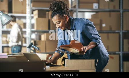 Besitzer eines kleinen Unternehmens, der einen stilvollen Fahrradsitz verpackt, der online an einen Kunden verkauft wird. Einen kleinen Karton für das Porto vorbereiten. Black Female Inventory Manager, der in der Lagereinrichtung arbeitet. Stockfoto