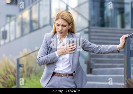 Eine blonde Frau in einem formellen grauen Anzug hält sich schmerzhaft an der Brust, während sie vor einem modernen Bürogebäude steht und einen Herzinfarkt oder starke Brustschmerzen erlebt. Stockfoto