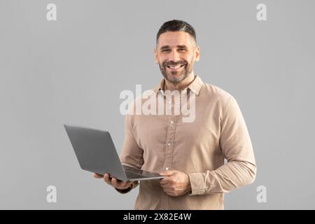 Mann hält Laptop und lächelt auf Grey Stockfoto
