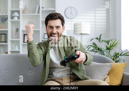Bärtiger Mann, der begeistert Videospiele auf einer Couch in einem stilvollen, gut beleuchteten Wohnzimmer spielt. Er feiert einen Sieg, während er einen Game-Controller hält. Stockfoto