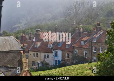 Ein malerischer Blick auf eine Reihe traditioneller englischer Cottages mit rot gekachelten Dächern und Kaminen vor einem nebeligen, grünen Hügel. Stockfoto