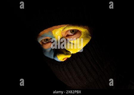 Das Gesicht eines Mädchens in einer schwarzen Sturmhaube und die Flagge der Ukraine sind im Dunkeln mit Farben auf ihr Gesicht gemalt Stockfoto