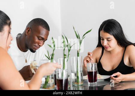 Drei junge Latino-Freunde, die im Speisesaal zu Mittag essen, während sie sich unterhalten, ein schwarzer Mann und zwei weiße Frauen Stockfoto