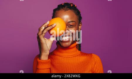 Lächelnde Afrikanerin mit verspieltem und lebendigem Stil steht in einem lila Studio. Sie hält eine Orangenfrucht, die einen gesunden Lebensstil zeigt Stockfoto