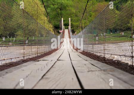 Fußgängerbrücke mit Metallzaun und Holzdielen, die über dem Fluss auf dem Hintergrund grüner Bäume hängen. Stockfoto