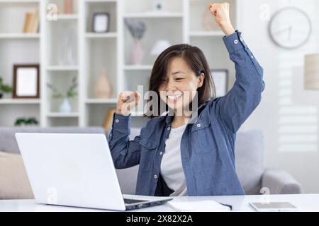 Die überfröhliche Frau hebt ihre Arme, während sie an einem Laptop arbeitet und zu Hause einen Moment des Sieges erlebt. Stockfoto