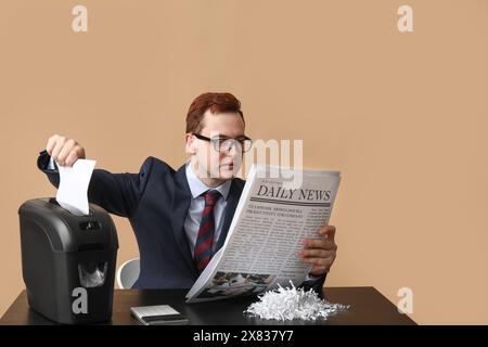 Hübscher junger Mann, der Zeitung liest und Papier im Aktenvernichter auf braunem Hintergrund zerstört Stockfoto