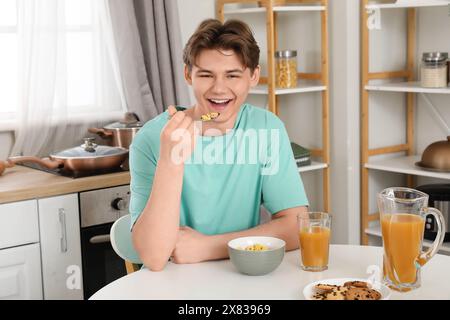 Ein glücklicher junger Mann isst Cornflakes mit Milch und trinkt Saft beim Frühstück zu Hause Stockfoto