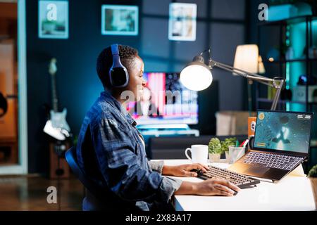 Afroamerikanerin spielt kompetitives Videospiel auf Laptop. Black Woman ist voll engagiert und in die Action eingetaucht, trägt Kopfhörer und nutzt den Bildschirm des Geräts, um das Gaming-Erlebnis zu verbessern. Stockfoto